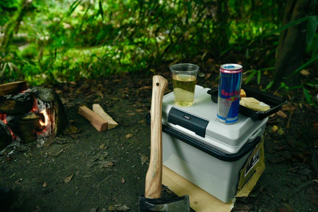 A serene campsite featuring an axe, fire, cooler, and energy drink for outdoor adventures.