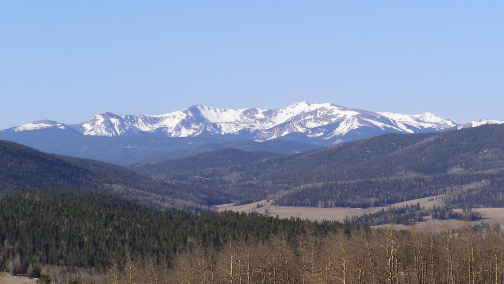 Scenic view of mountains rising above a lush field dotted with trees under a clear blue sky.