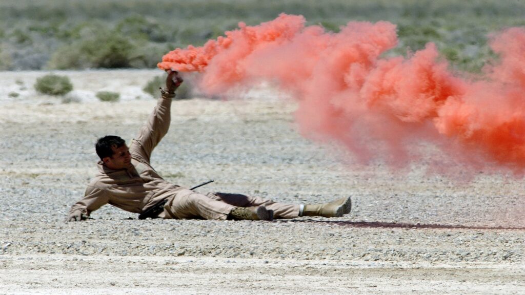 A man sprawls on the ground, with a red smoke bomb releasing a vivid plume, enhancing the atmosphere of the moment.