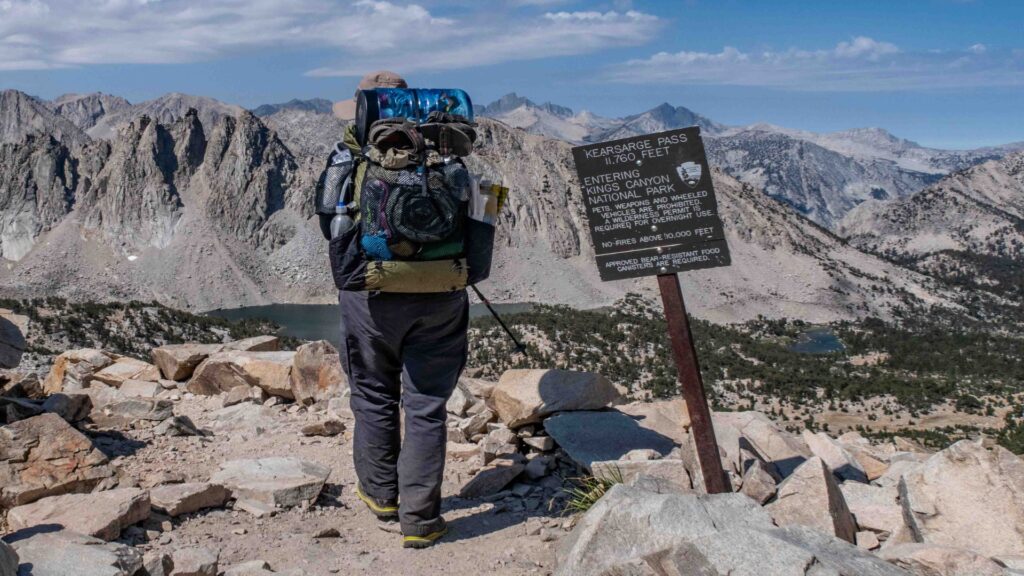 A hiker stands triumphantly on a mountain peak, backpack on, overlooking a vast landscape of valleys and distant peaks.