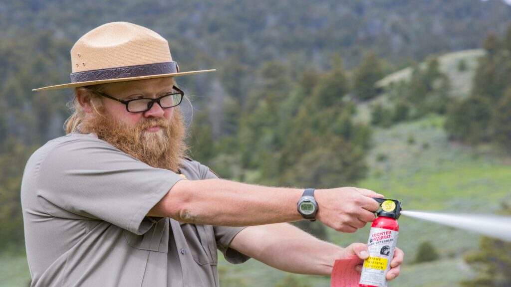 A man with a beard and hat uses bear spray demonstrating safety in a wilderness setting.