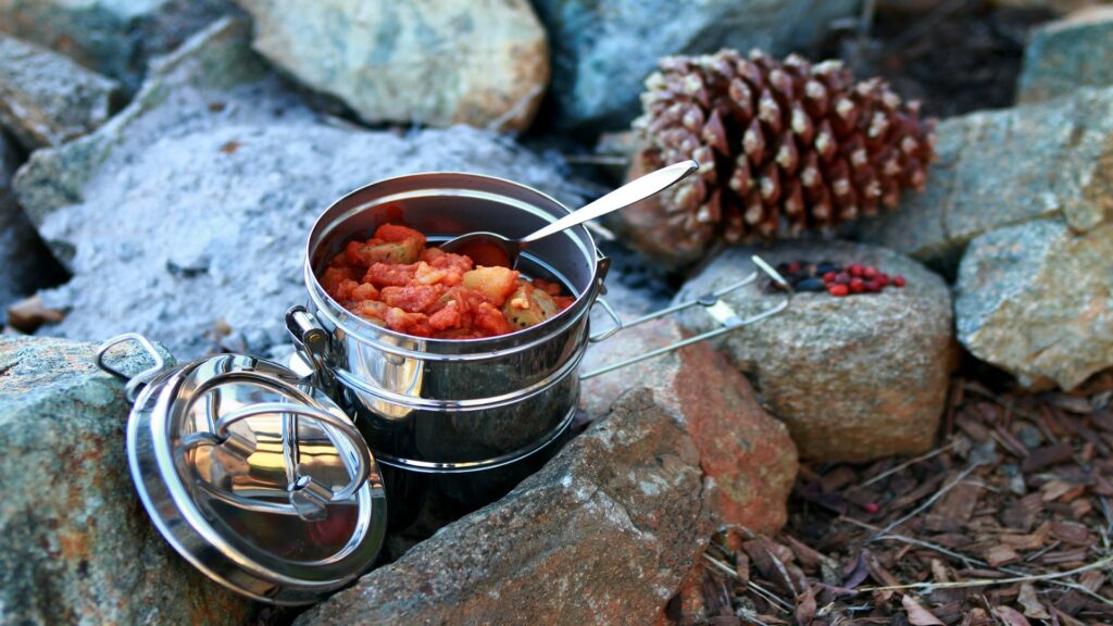 A metal container with a spoon rests on rocks, filled with food, suggesting a rustic outdoor dining experience.
