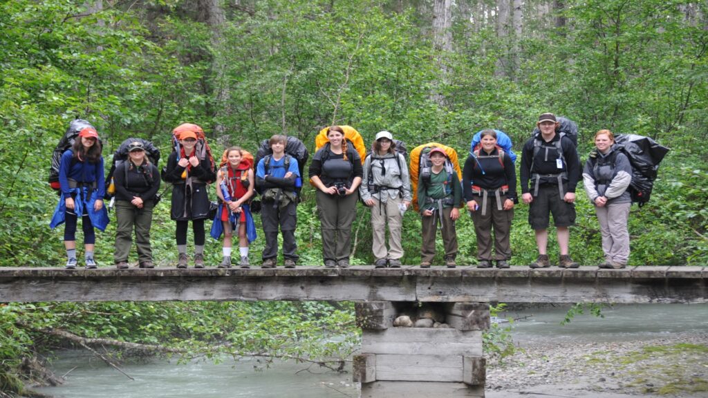 A diverse group of people with backpacks stands on a bridge, capturing the moment together against a scenic backdrop.