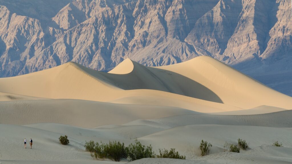 Two hikers traverse golden sand dunes with rugged mountains rising in the background at Death Valley National Park.