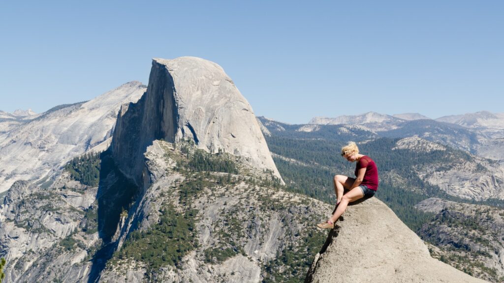 A woman sits on a rock, gazing at a majestic mountain landscape under a clear blue sky.