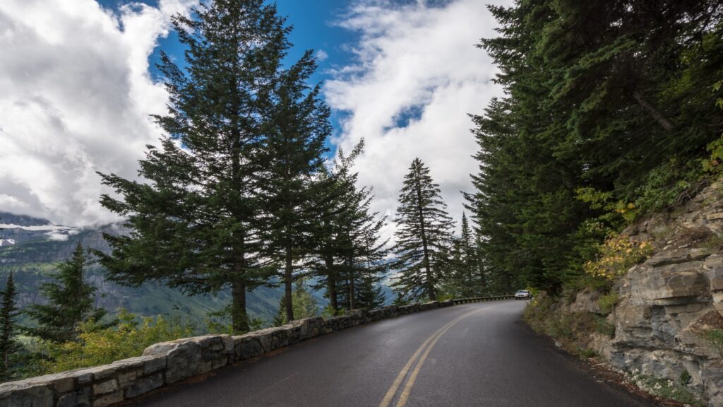 A scenic roadway flanked by vibrant trees, with stunning mountains looming in the background under a sunny sky.