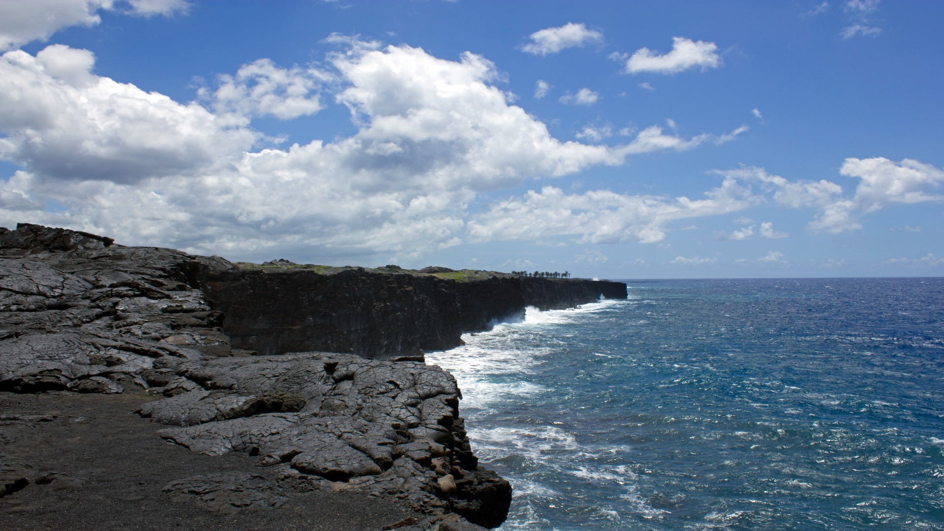 Expansive view of Hawai'i Volcanoes National Park reflecting the sky, surrounded by lush greenery and distant hills.