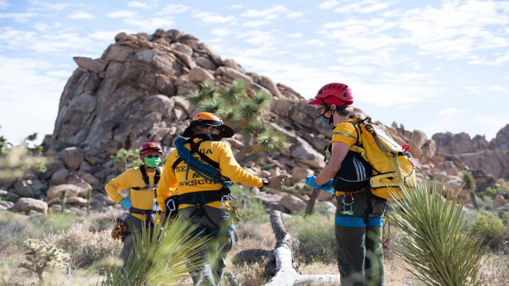 Three rescuers in yellow jackets stand in a vast desert, ready to assist those in need under the bright sun.