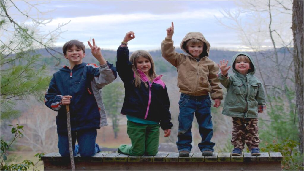 Four children joyfully standing on a wooden bench, raising their hands in excitement against a bright outdoor backdrop.