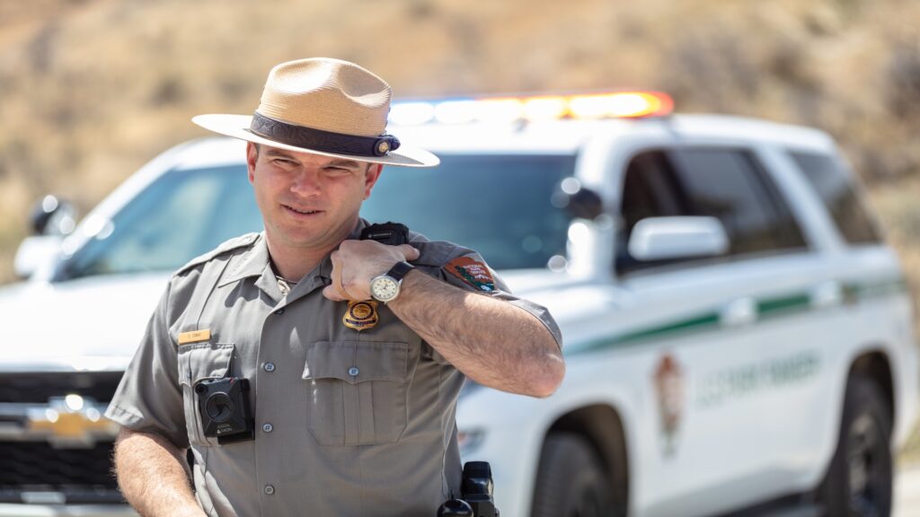 A man in a hat stands by a police car, using a radio to coordinate efforts in a law enforcement operation.