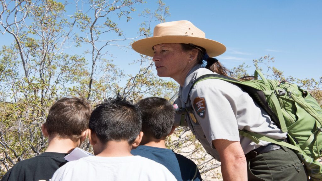 A park ranger in a hat engages with children, sharing knowledge about nature in a vibrant outdoor setting.