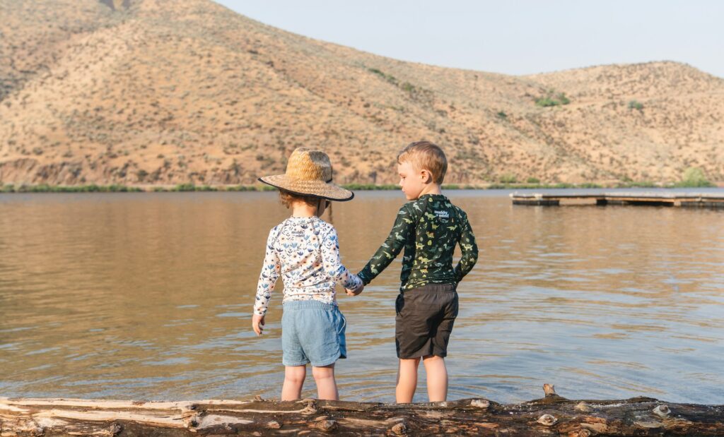Two kids stand on a log in the water, surrounded by nature, sharing a moment of fun and exploration.