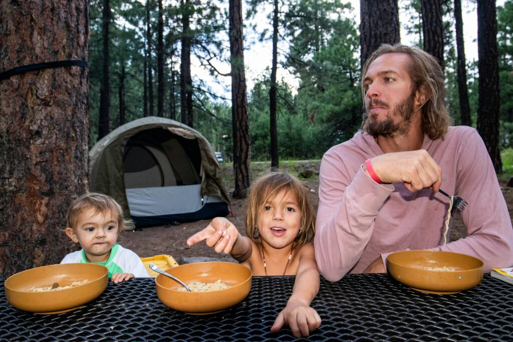 A father and his two kids sit at a campground, cheerfully eating cereal from bowls while enjoying the outdoors.