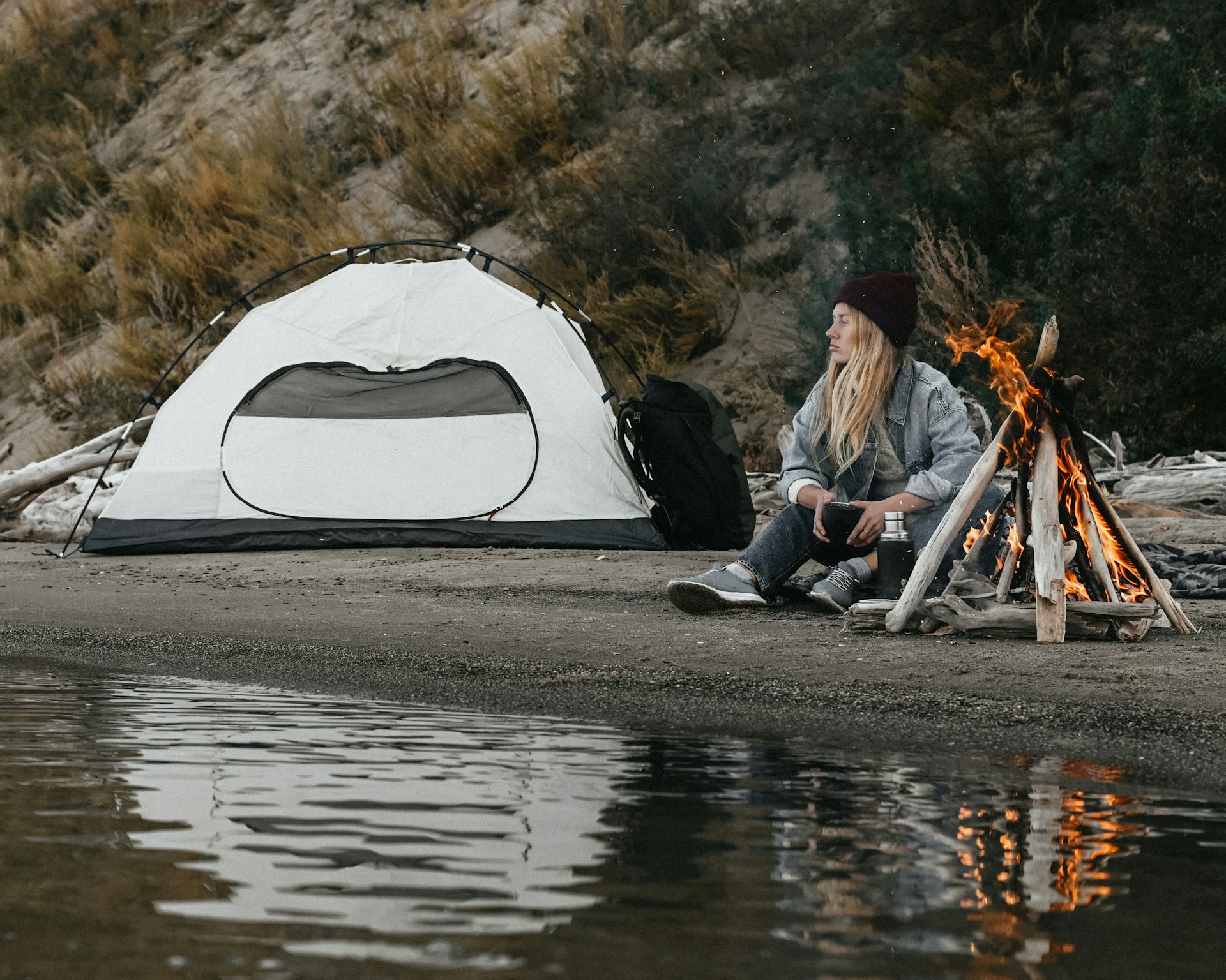 A woman enjoys the warmth of a campfire, seated next to her tent in a peaceful camping scene.