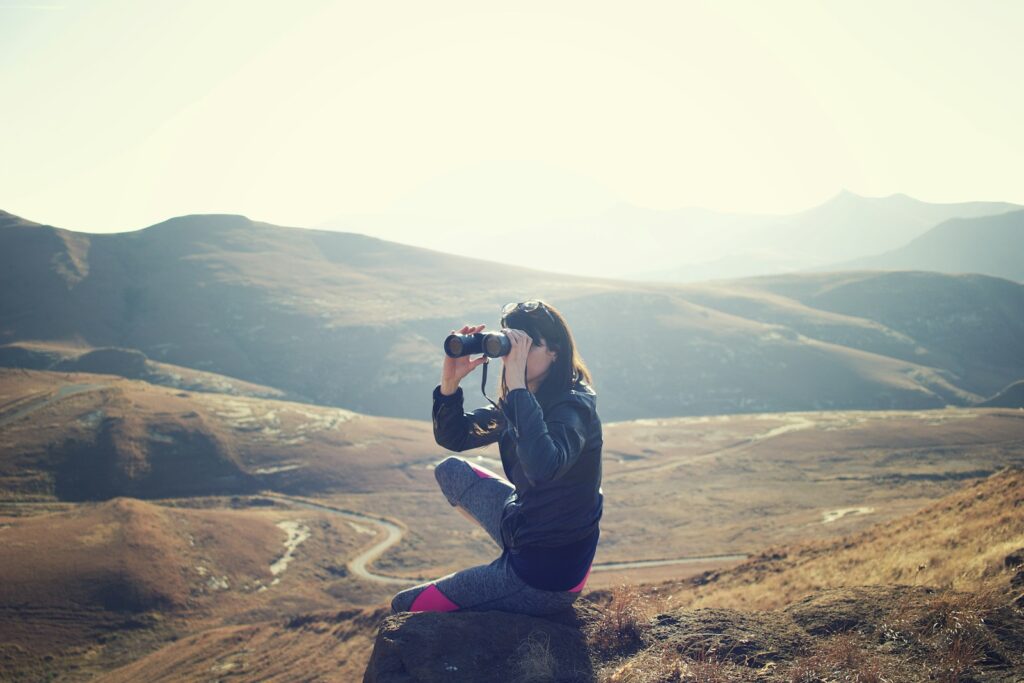 Woman wearing black jacket sitting on gray rock and using binoculars.