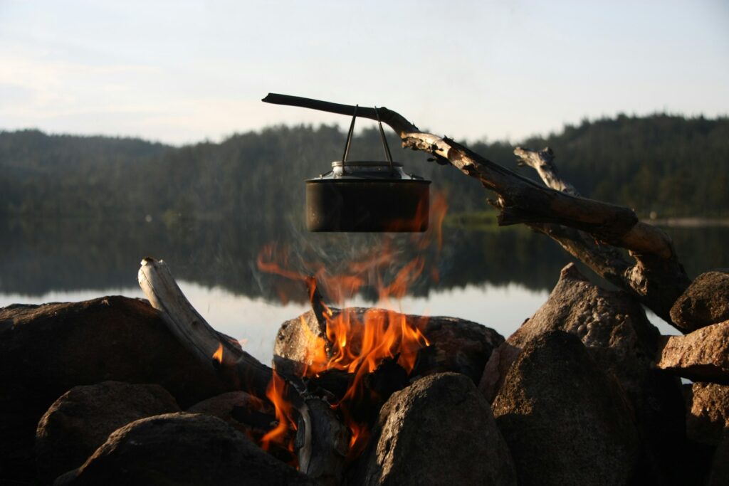 A pot sits over a fire, framed by a tranquil lake, showcasing a peaceful outdoor cooking scene.