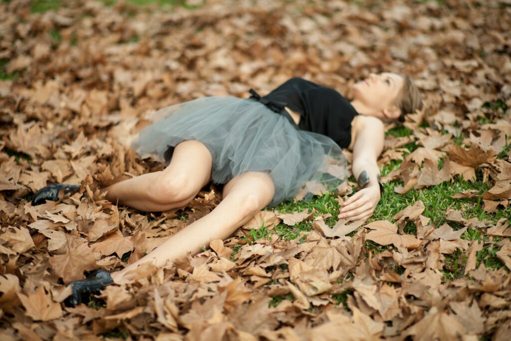 A woman lies on the ground covered in fallen leaves, immersed in the beauty of nature during autumn.