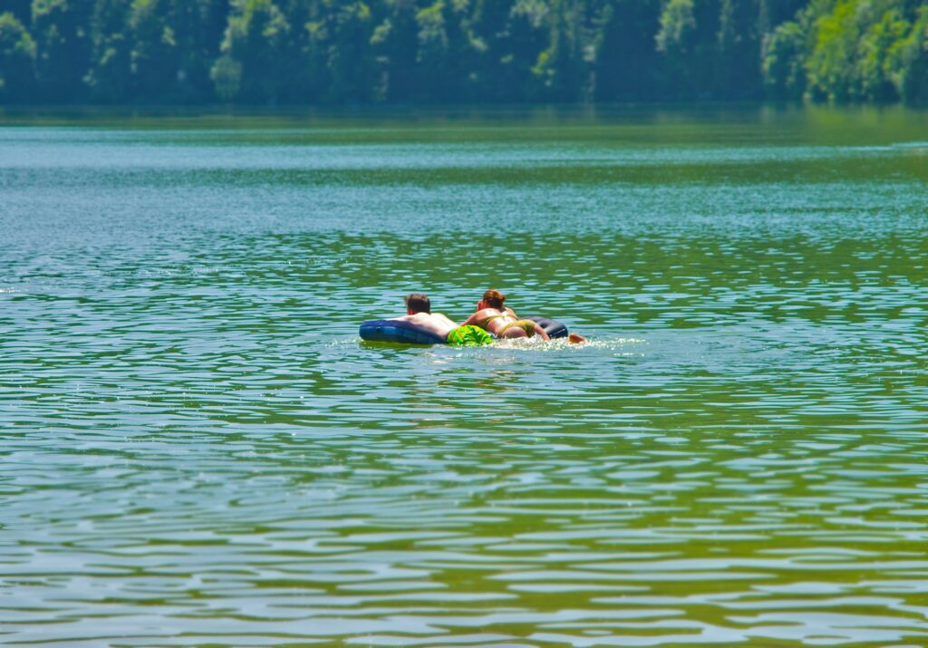 A pair of friends enjoy a sunny day, drifting on an inner tube in a calm lake surrounded by lush greenery.