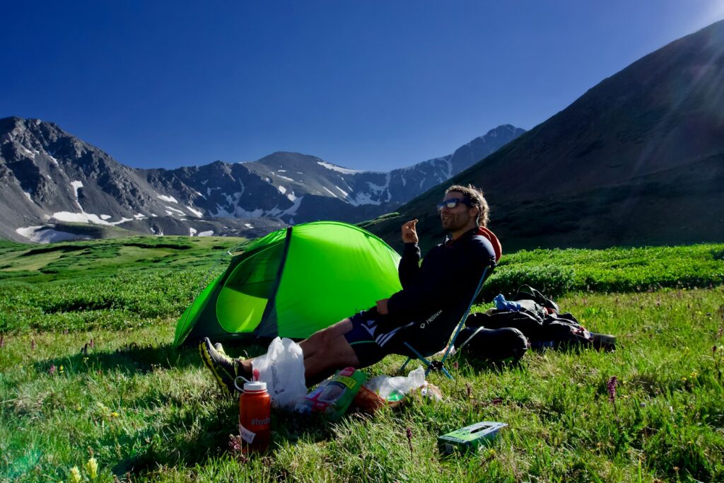 Man sitting on black camping chair beside green dome tent.
