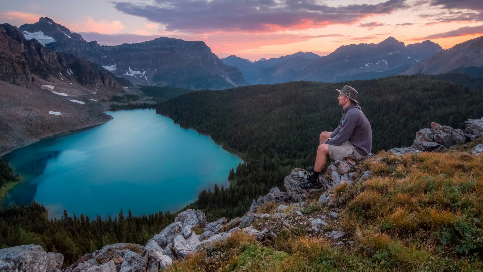 A man relaxes on a mountain summit, overlooking a peaceful lake, with expansive views of nature stretching beyond.