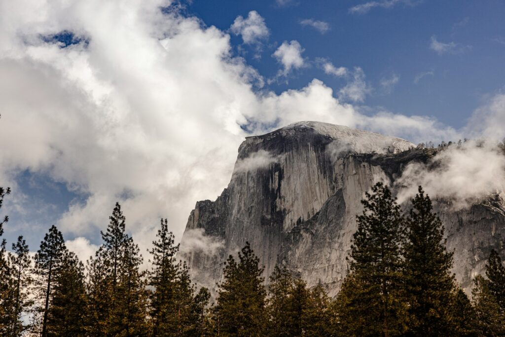 A majestic mountain rises behind a foreground of lush trees, partially shrouded by soft, white clouds.