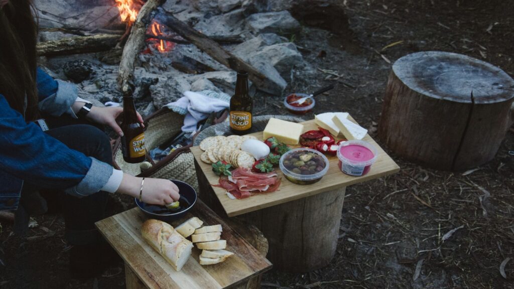 A person relaxes by a campfire, enjoying food and drinks under a starlit sky, creating a cozy outdoor atmosphere.