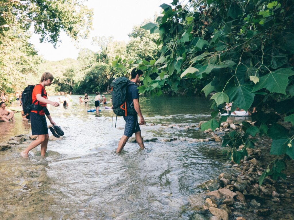 Two backpackers traverse a river, stepping over rocks and water, immersed in a vibrant outdoor landscape.