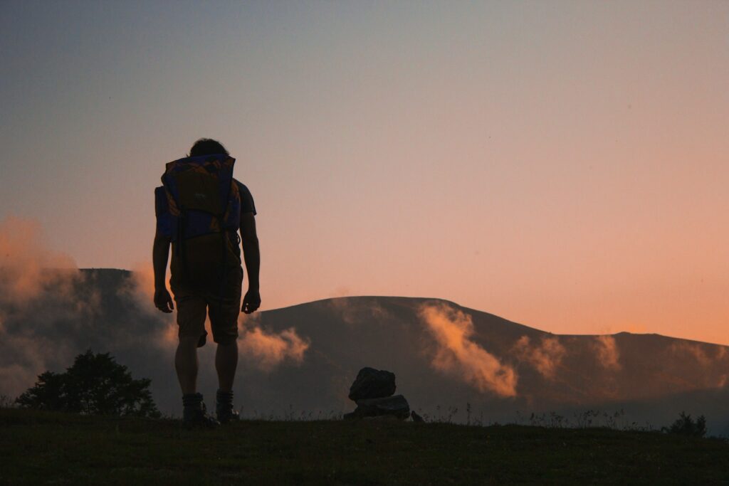 A hiker with a backpack stands on a hill, ready to ignite a fire with mountains rising in the background.