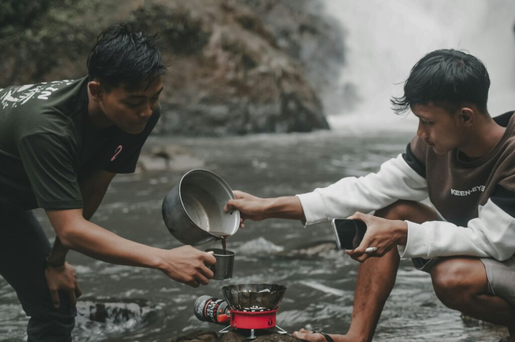 Two men prepare food on a stove near a cascading waterfall, showcasing a unique outdoor cooking experience.