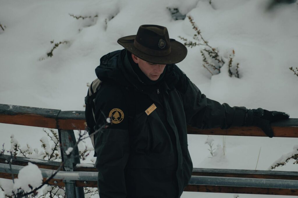 A park ranger in winter attire stands on a snow-covered bridge, reflecting tranquility in nature.