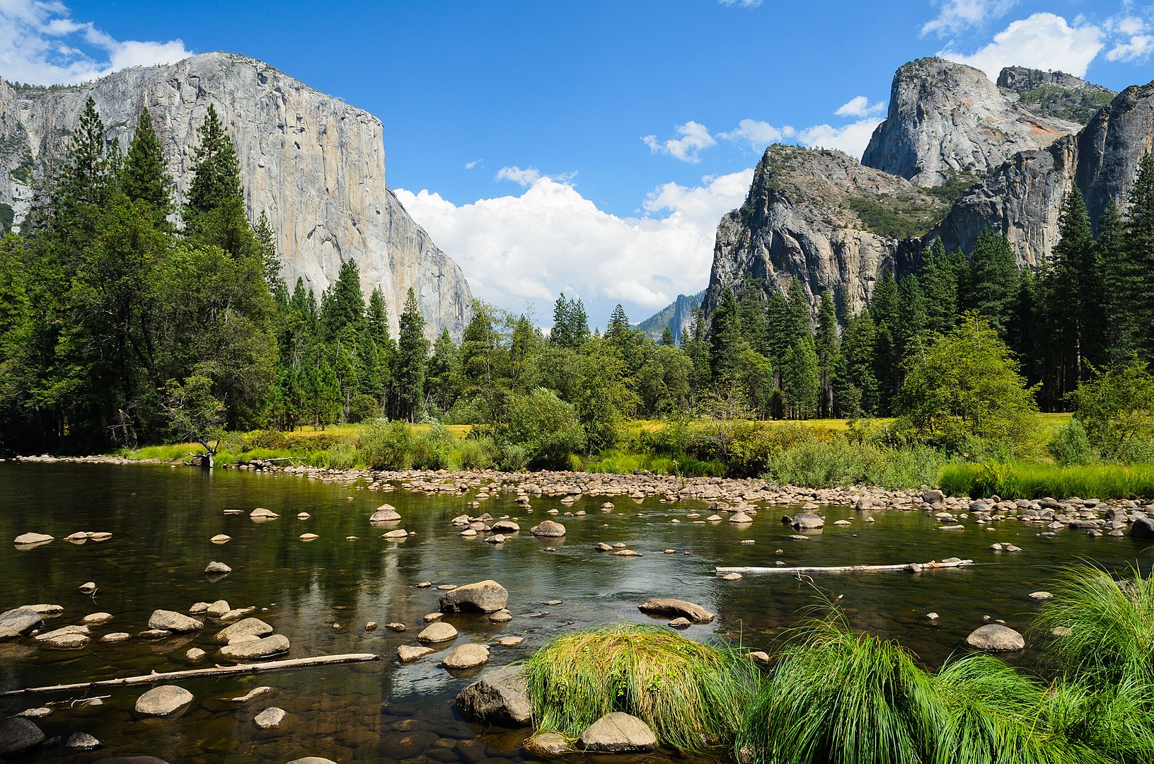 A serene landscape featuring towering granite cliffs, lush green trees, and a calm river dotted with rocks under a blue sky.