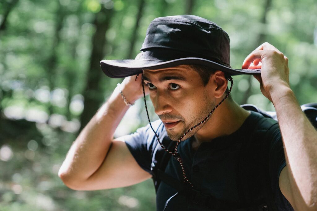 A young man adjusts his hat while hiking through a sunlit forest, enjoying modern outdoor adventure.