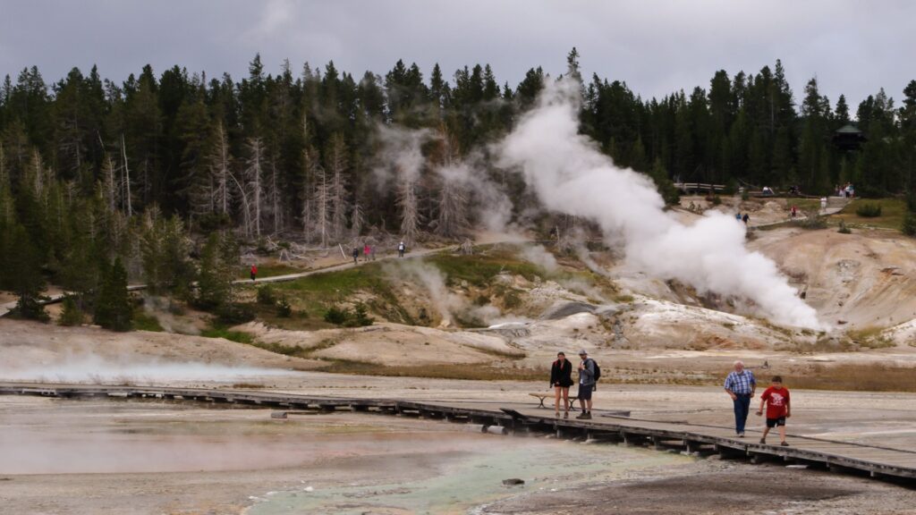 A family explores a wooden boardwalk near a geyser, experiencing the unique geothermal wonders of Yellowstone National Park.