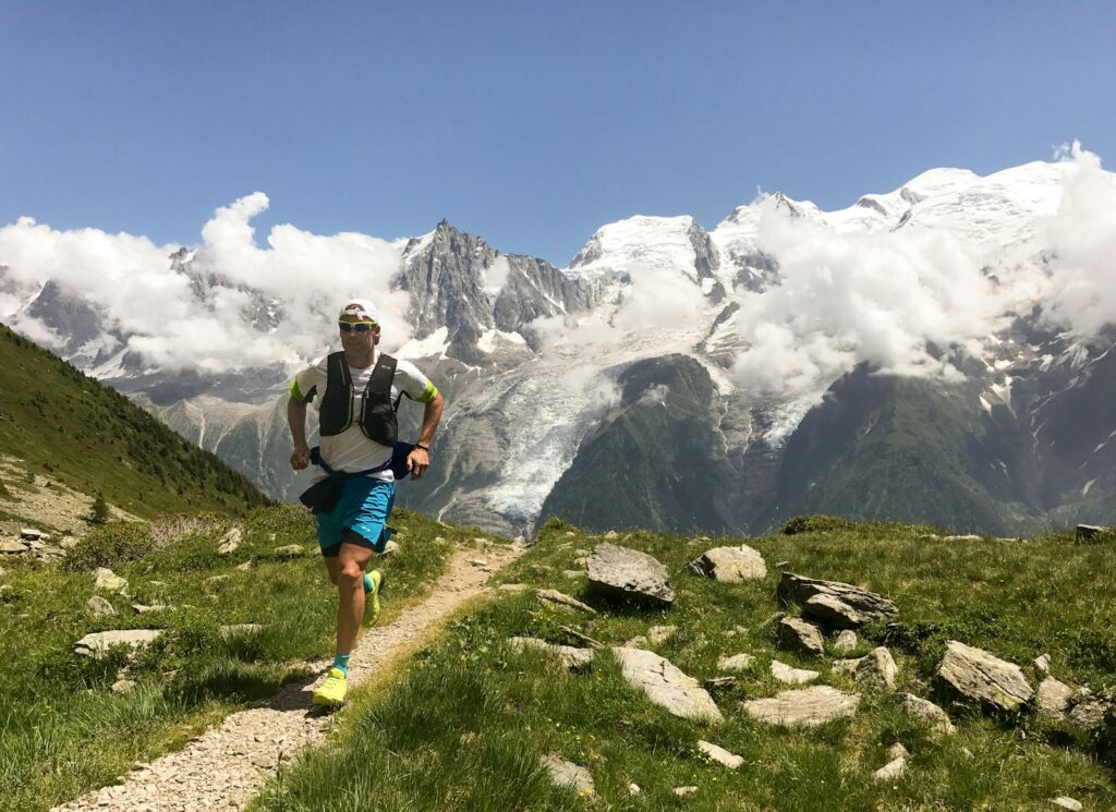 Man in white shirt and blue shorts running on brown ground between green grass during daytime.