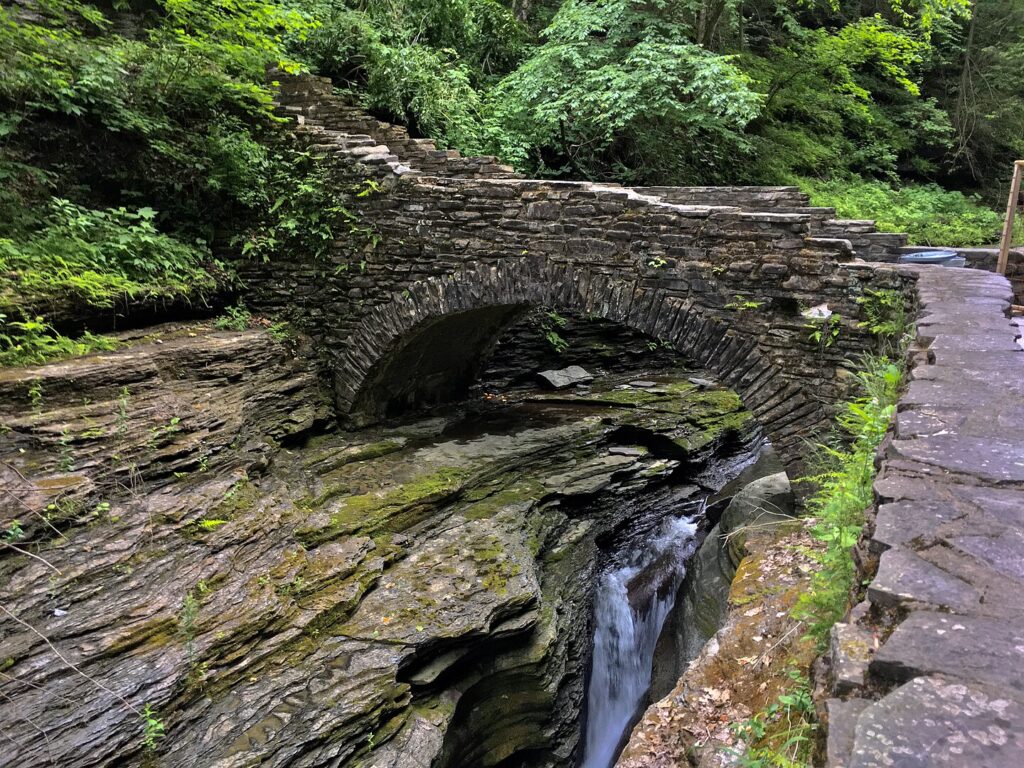 A stone bridge arches over a serene stream, surrounded by lush woods in Watkins Glen State Park.